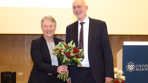 Zwei lächelnde Personen stehen nebeneinander auf einer Bühne und tragen schicke Kleidung. Die Frau, Barbara Windscheid, hält einen Blumenstrauß in ihren Händen. Neben ihr steht Prof. Dr. Thomas Puhl.