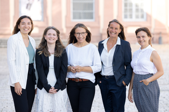Fünf lächelnde Frauen stehen in einer Reihe vor den historischen Gebäuden im Ehrenhof des Mannheimer Schlosses.