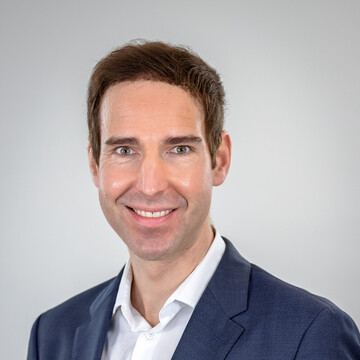 Prof. Dr. Florian Weigert. He is wearing a white shirt with a blue jacket over it and is standing in front of a white wall.