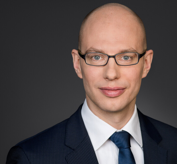 Portrait von Nils Schaks. Er trägt ein weißes Hemd, eine dunkelblaue Krawatte sowie einen dunkelblauen Blazer und steht vor einer dunkelgrauen Wand. 