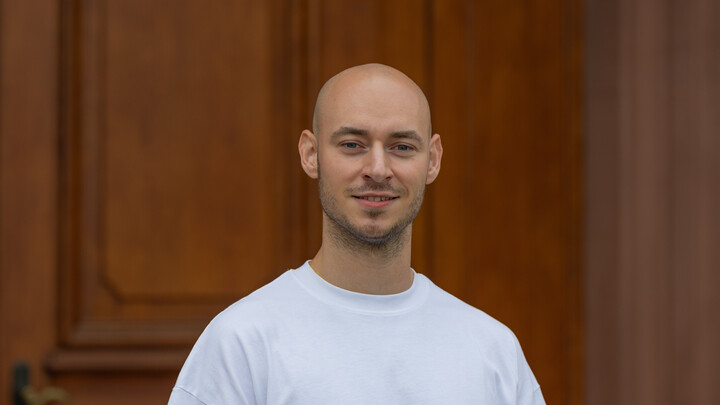 Maximilian Volmer. He is wearing a white T-shirt and is standing in front of a brown door.
