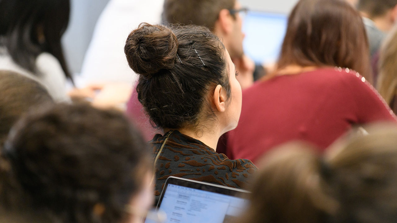 Eine Studentin hört aufmerksam einem Vortrag zu. Sie ist von hinten zu sehen, hat dunkelbraune Haare und trägt einen Dutt.