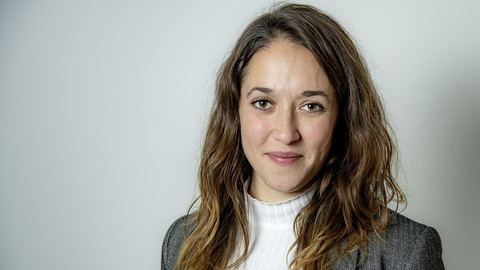 Matilde Cappeletti. She is wearing a grey jacket over a white sweater and is standing in front of a grey wall.