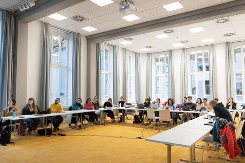 Eine Gruppe von Menschen sitzt an Tischen, die u-förmig angeordnet sind. Einige schreiben etwas während viele nach vorne blicken.