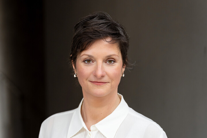 Frau Schäfer trägt eine weiße Bluse. Sie steht vor einem dunklen Hintergrund.