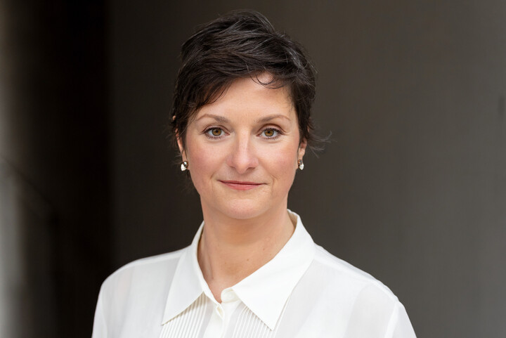 Frau Schäfer trägt eine weiße Bluse. Sie steht vor einem dunklen Hintergrund.