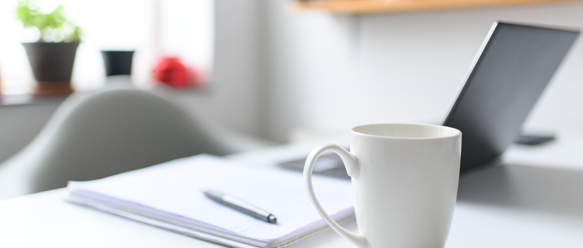 Eine Tasse steht auf einem Tisch. Im Hintergrund liegen ein Notizblock und ein Laptop.