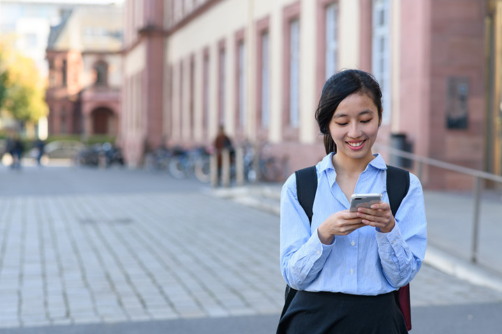 Eine Studentin steht lächelnd vor dem Schloss der Universität Mannheim und schaut auf ihr Handy.