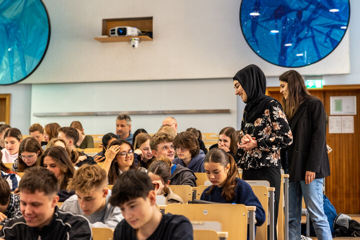 Jugendliche sitzen in einem Hörsaal. Zwei Dozentinnen stehen neben ihren Sitzen und erklären ihnen etwas.