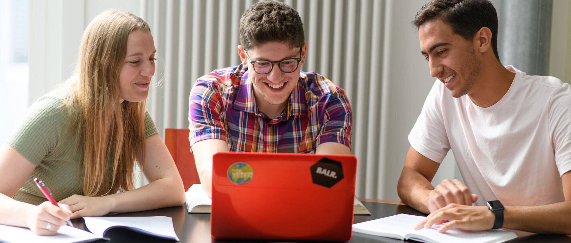 Drei Studierende sitzen mit ihren Arbeitsmaterialien vor einem roten Laptop