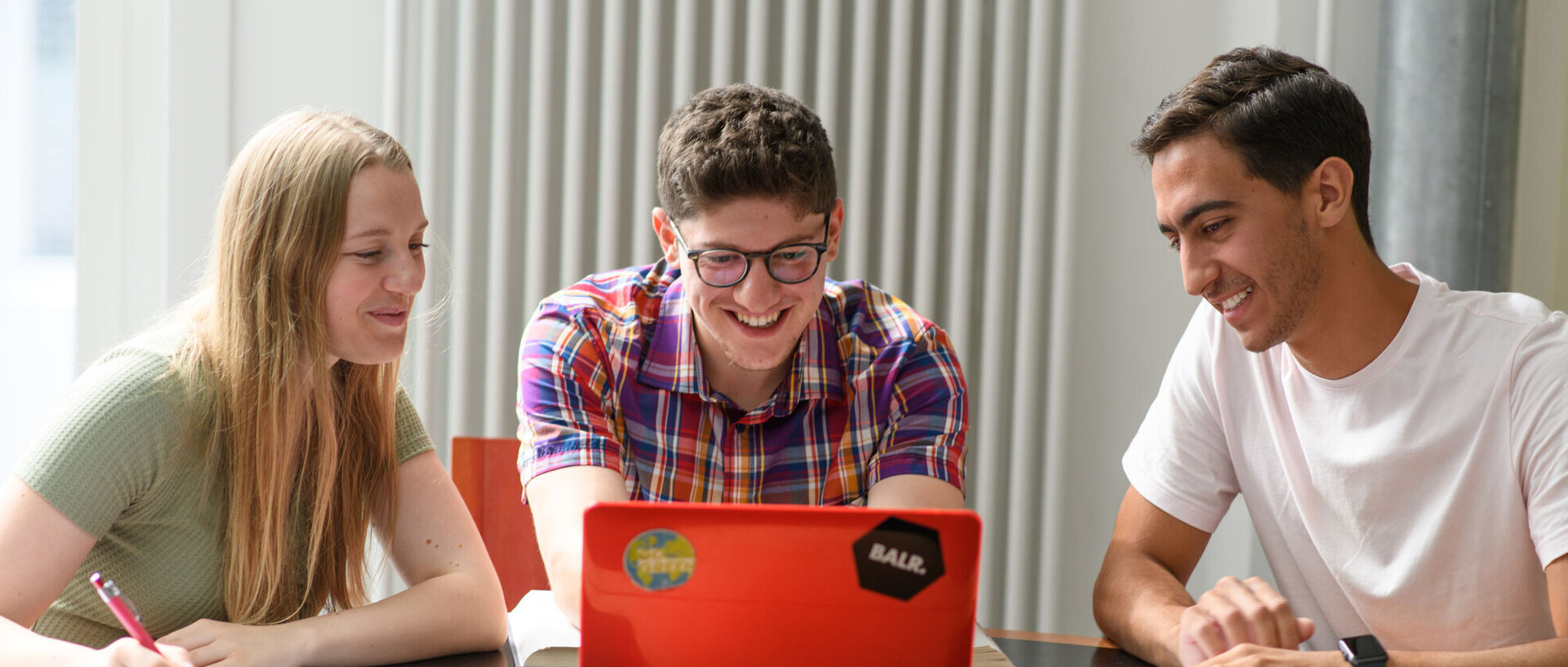 Drei Studierende sitzen mit ihren Arbeitsmaterialien vor einem roten Laptop