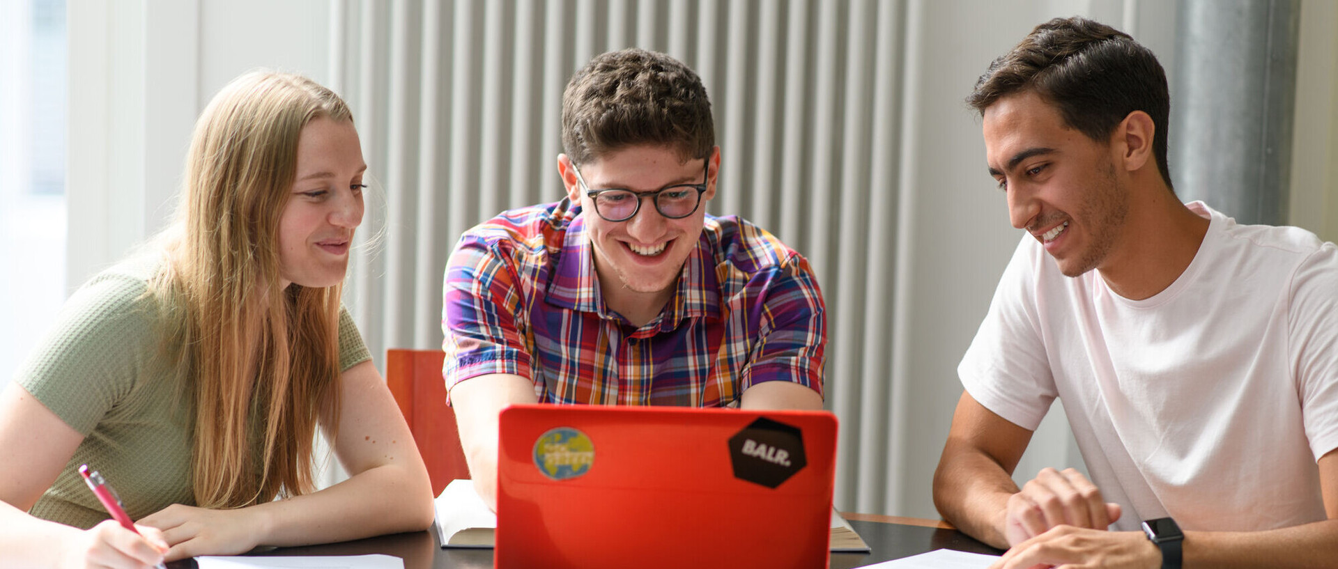 Drei Studierende sitzen mit ihren Arbeitsmaterialien vor einem roten Laptop