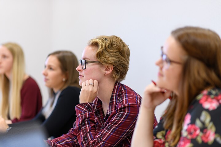 Studierende sitzen an einem Tisch und hören bei einem Vortrag zu.