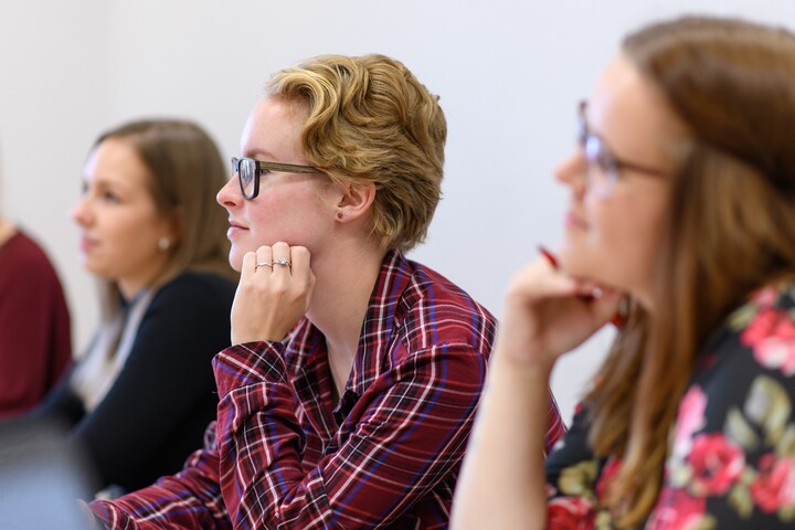 Studierende sitzen an einem Tisch und hören bei einem Vortrag zu.