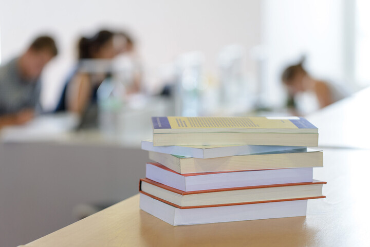 Ein Bücherstapel liegt auf einem Tisch. Im Hintergrund die Umrisse mehrerer Personen an einem Tisch. 