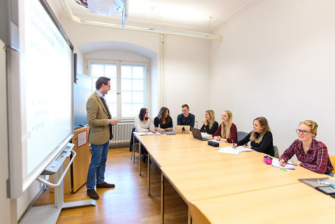 Ein Dozent unterhält sich in einem Seminarraum mit einer kleinen Gruppe aus Studierenden.