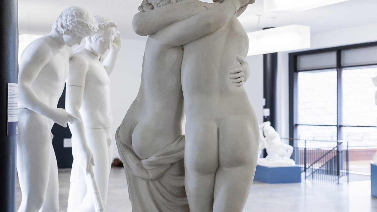 In einem Gang sind antike Skulpturen ausgestellt. Im Mittelpunkt steht eine Skulptur von einer halbnackten und einer nackten Figur, die einander mit dem Rücken zur Kamera in den Armen halten.