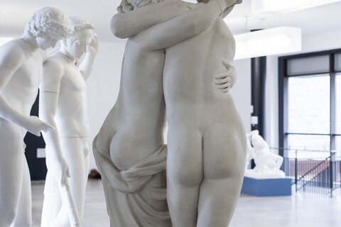 In einem Gang sind antike Skulpturen ausgestellt. Im Mittelpunkt steht eine Skulptur von einer halbnackten und einer nackten Figur, die einander mit dem Rücken zur Kamera in den Armen halten.