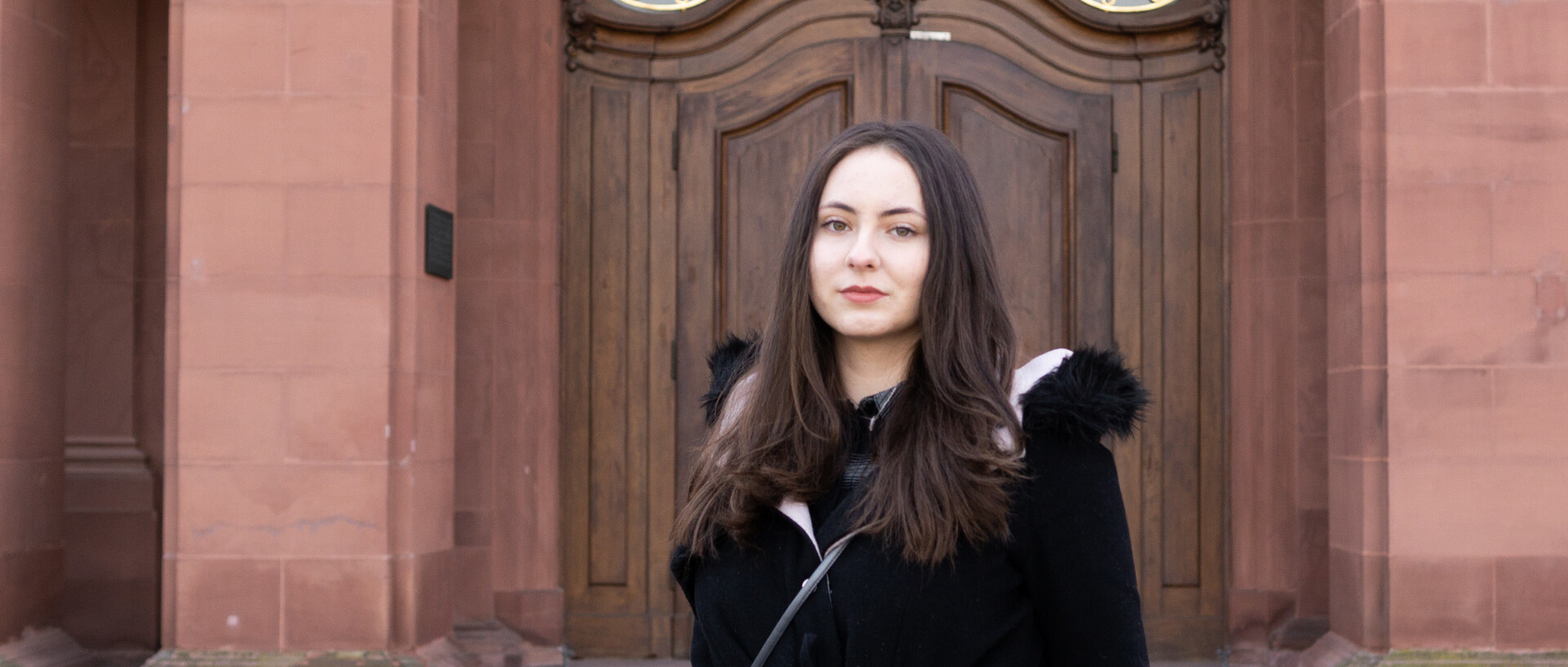 Eine Person trägt eine schwarze Jacke mit schwarzem Fell und steht vor einer braunen Schlosstüre. Die Person heißt Dijana Babic.