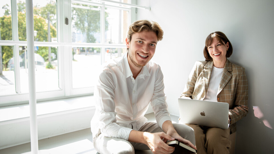 Zwei Forschende sitzen auf einer Fensterbank. Die Frau arbeitet an einem Laptop. Der Mann hält ein Notizbuch in den Händen. Beide lachen. 