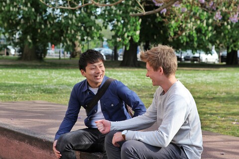 Zwei Studenten sitzen auf Steinen und lachen gemeinsam. Im Hintergrund blühen ein paar Bäume.