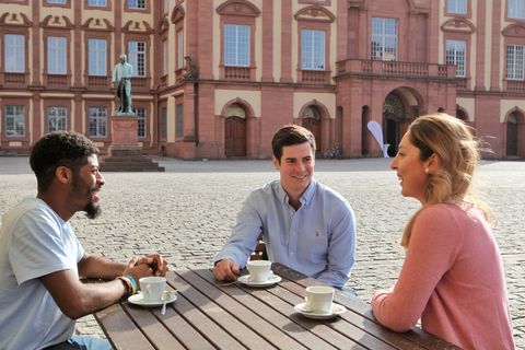 Zwei junge Männer und eine junge Frau sitzen an einem Holztisch auf dem Ehrenhof. Vor ihnen stehen drei weiße Tassen.