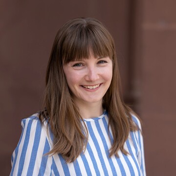Caterina Katzer hat braune lange Haare und ein Pony. Sie trägt ein blau-weiß-gestreiftes Oberteil.