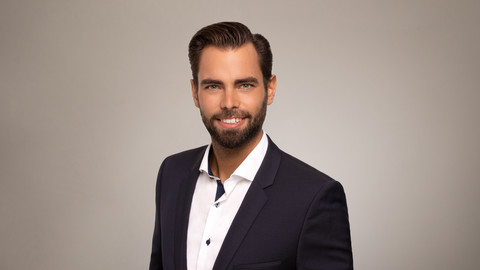 Frank Ohnesorge. He is wearing a black suit and is standing in front of a brownish photowall.