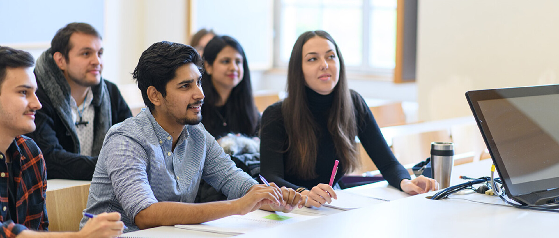 Studierende sitzen in einem Seminarraum an Tischen und blicken nach vorne. Auf den Tischen liegen Notizblöcke und Mäppchen.
