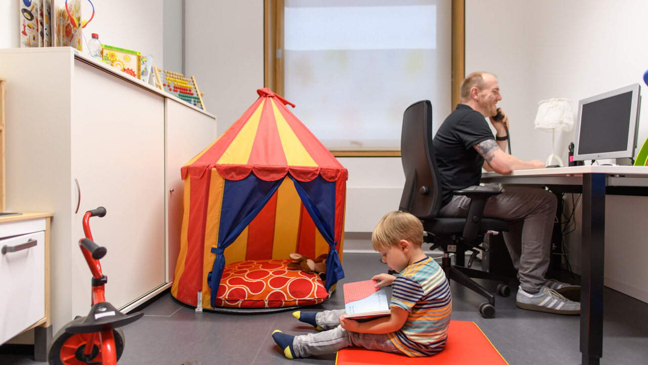 Kind sitzt in einem Spielraum und betrachtet ein Buch, ein Mann beaufsichtigt es.