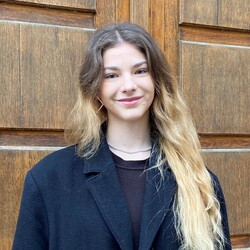 Helen Kalmbach is standing in front of a round wooden door at the University of Mannheim. She is wearing a long black wool coat and her long hair is worn loose.