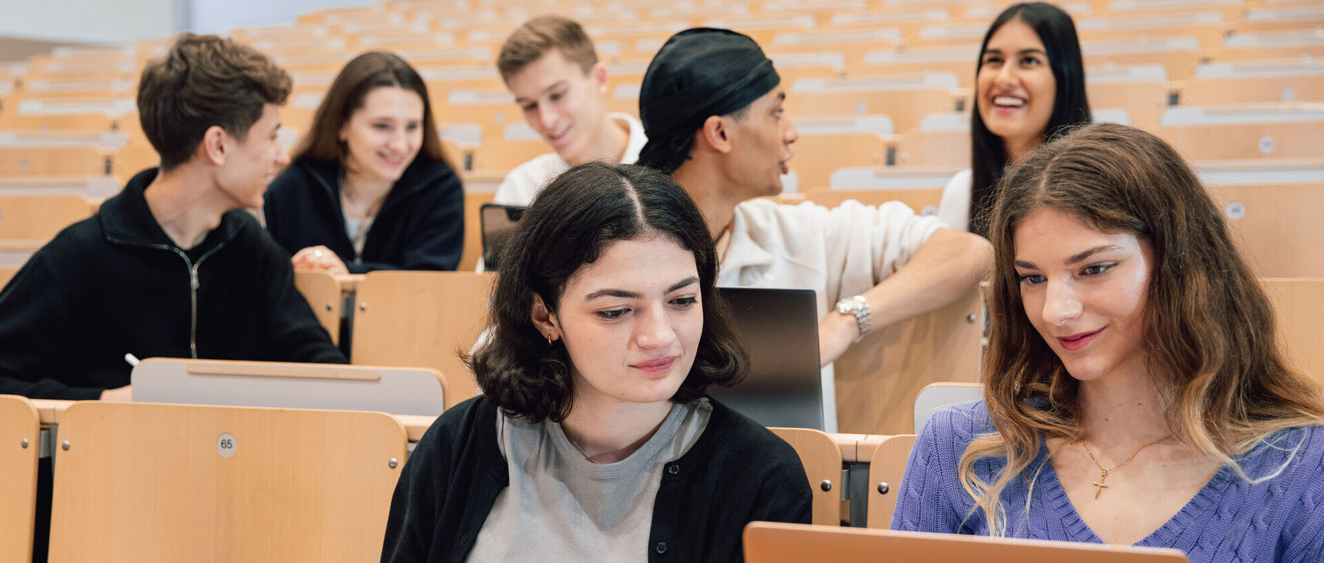 Eine Gruppe von Studierenden sitzen in einem Hörsaal uns sprechen miteinander. Zwei Studentinnen betrachten etwas an einem aufgeklappten Laptop.