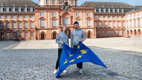 Zwei Personen tragen einen grauen Pullover der Uni Mannheim und halten die Fahne der Europäischen Union in ihren Händen. Sie stehen auf dem Ehrenhof.