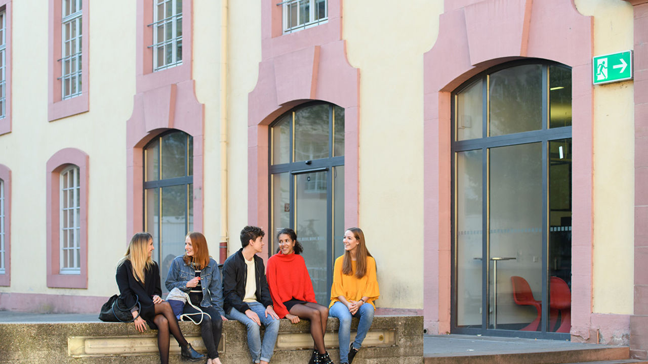 Fünf Studierende mit bunten Pullovern und Jeansjacke sitzen lachend auf einer Steinmauer im Schneckenhof