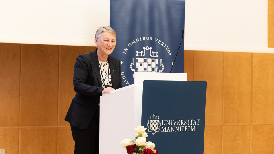 Portrait von Barbara Windscheid. Sie trägt eine weiße Bluse und einen schwarzen Blazer und steht an einem Rednerpult.