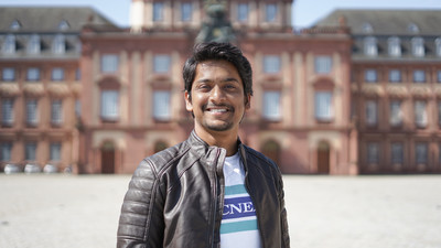 Eine lächelnde Person trägt ein weißes T-Shirt mit hell- und dunkelblauen Streifen und steht auf dem Ehrenhof vor dem Schloss. Die Person heißt Afham Uddin Sheikh.