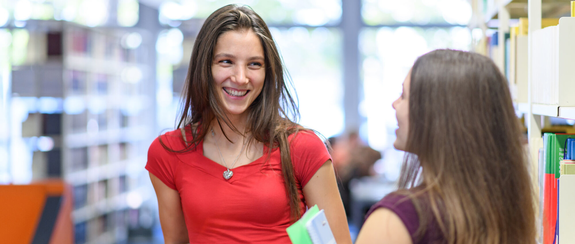 Zwei Studentinnen unterhalten sich stehend in der Bibliothek