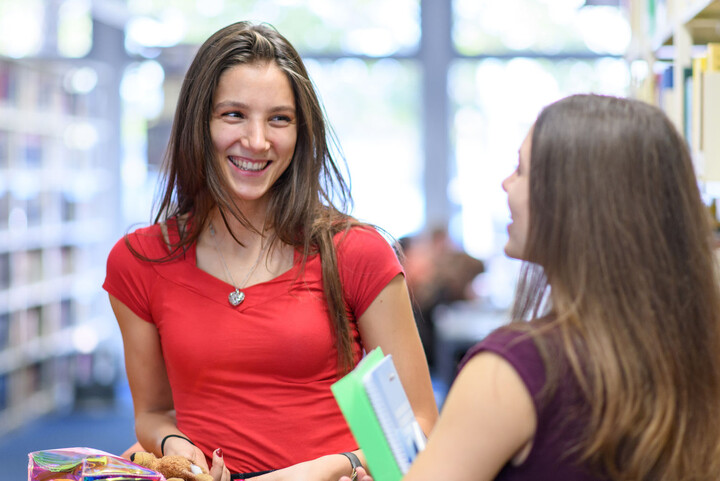 Two students are standing opposite each other and talking in the library.