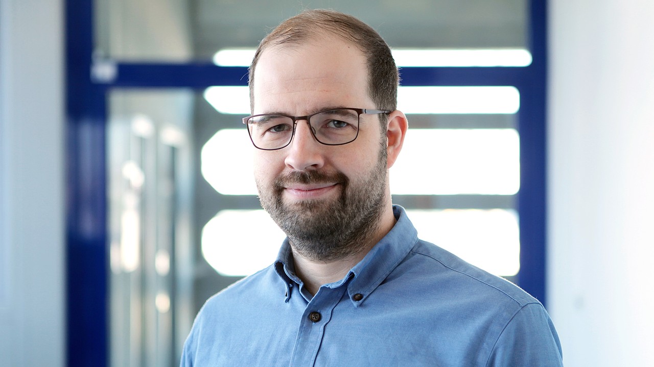 Dr. Lars Leszczensky hat einen Bart und kurze braune Haare. Er trägt ein blaues Hemd.