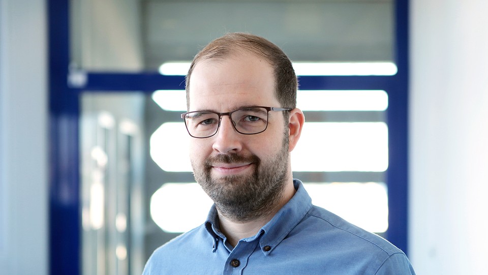 Dr. Lars Leszczensky hat einen Bart und kurze braune Haare. Er trägt ein blaues Hemd.