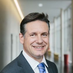 Alexander Schwörer hat dunkelbraune, leicht graue Haare und braune Augen. Er lächelt und trägt ein weißes Hemd, ein dunkelgraues Sakko und eine blau, weiß gestreifte Kravatte.