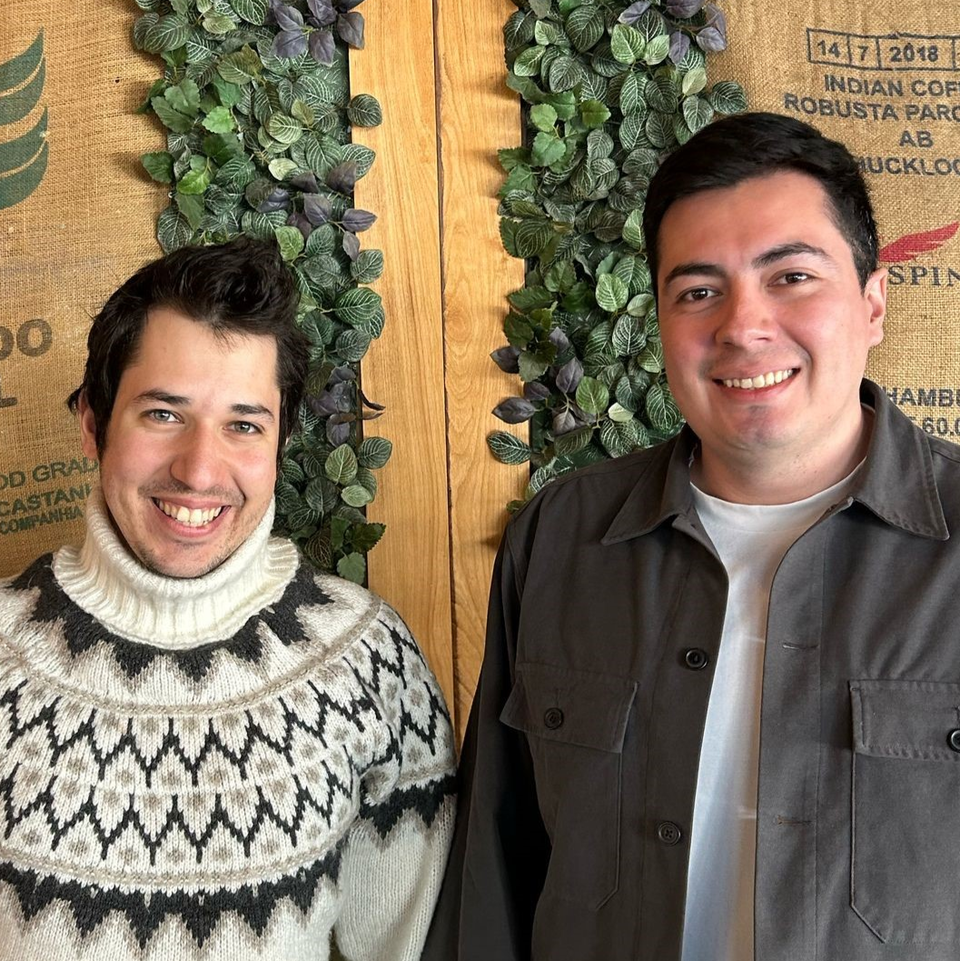 Pablo und Taylan stehen vor einer mit Blätterranken verkleideten Holzwand. Sie lachen in die Kamera.