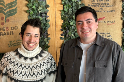 Pablo und Taylan stehen vor einer mit Blätterranken verkleideten Holzwand. Sie lachen in die Kamera.
