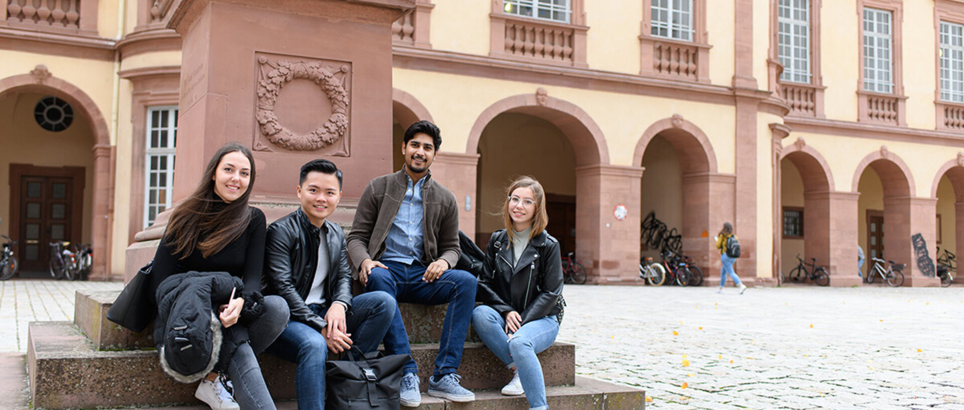 Zwei Studentinnen und zwei Studenten sitzen auf dem Ehrenhof der Universität Mannheim vor einer Statue.