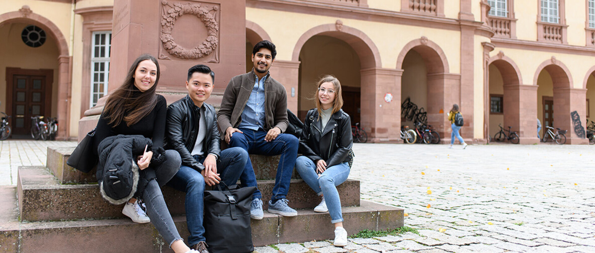 Zwei Studentinnen und zwei Studenten sitzen auf dem Ehrenhof der Universität Mannheim vor einer Statue.