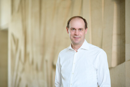 Eine Personn trägt ein weißes Hemd und steht vor einer beige-farbenen Wand. Die Person heißt Jannis Bischof.