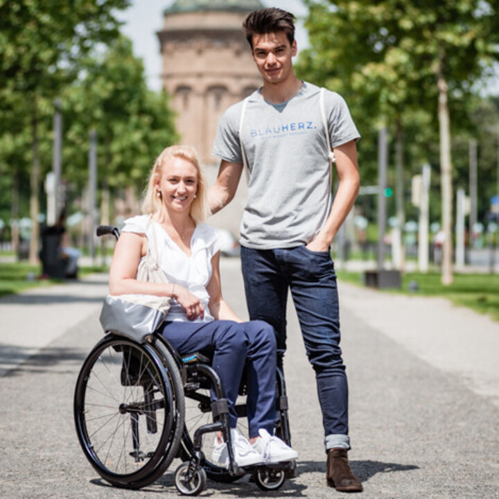 Eine Frau sitzt in einem Rollstuhl vor dem Wasserturm. Neben ihr steht ein Mann.