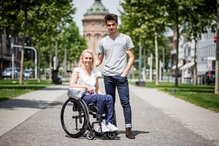 Eine Frau sitzt in einem Rollstuhl vor dem Wasserturm. Neben ihr steht ein Mann.