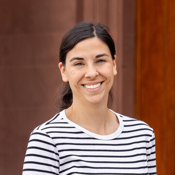 Eva Breitbach has long brown hair tied back in a ponytail and is wearing a white and blue striped long-sleeved shirt. She is shown smiling in front of Mannheim Palace..
