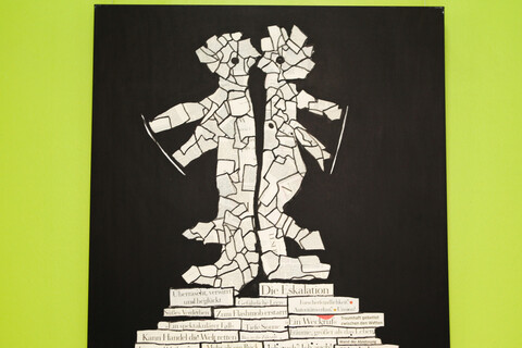 Vor einer grünen Wand hängt ein Kunstwerk. Abgebildet ist eine Zeitungscollage in Form von zwei Figuren vor schwarzem Hintergrund.